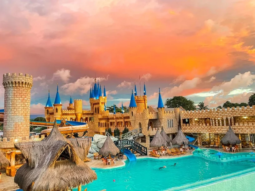 Castillo de piedra con torres de techo azul junto a una piscina con toboganes y kioscos de paja, al atardecer